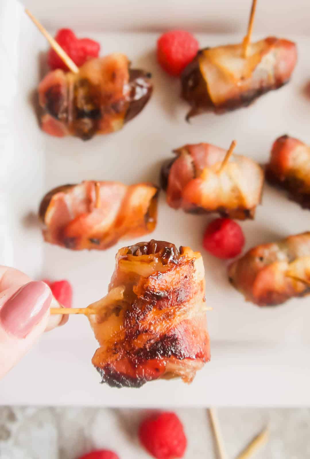 Raspberry stuffed medjool date covered in bacon being lifted by a tooth pick.