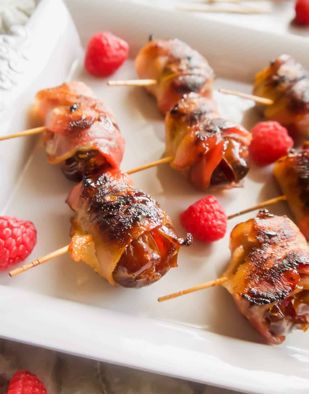 Rasberry stuffed Dates Wrapped in Bacon on a plate with raspberries surrounding them.