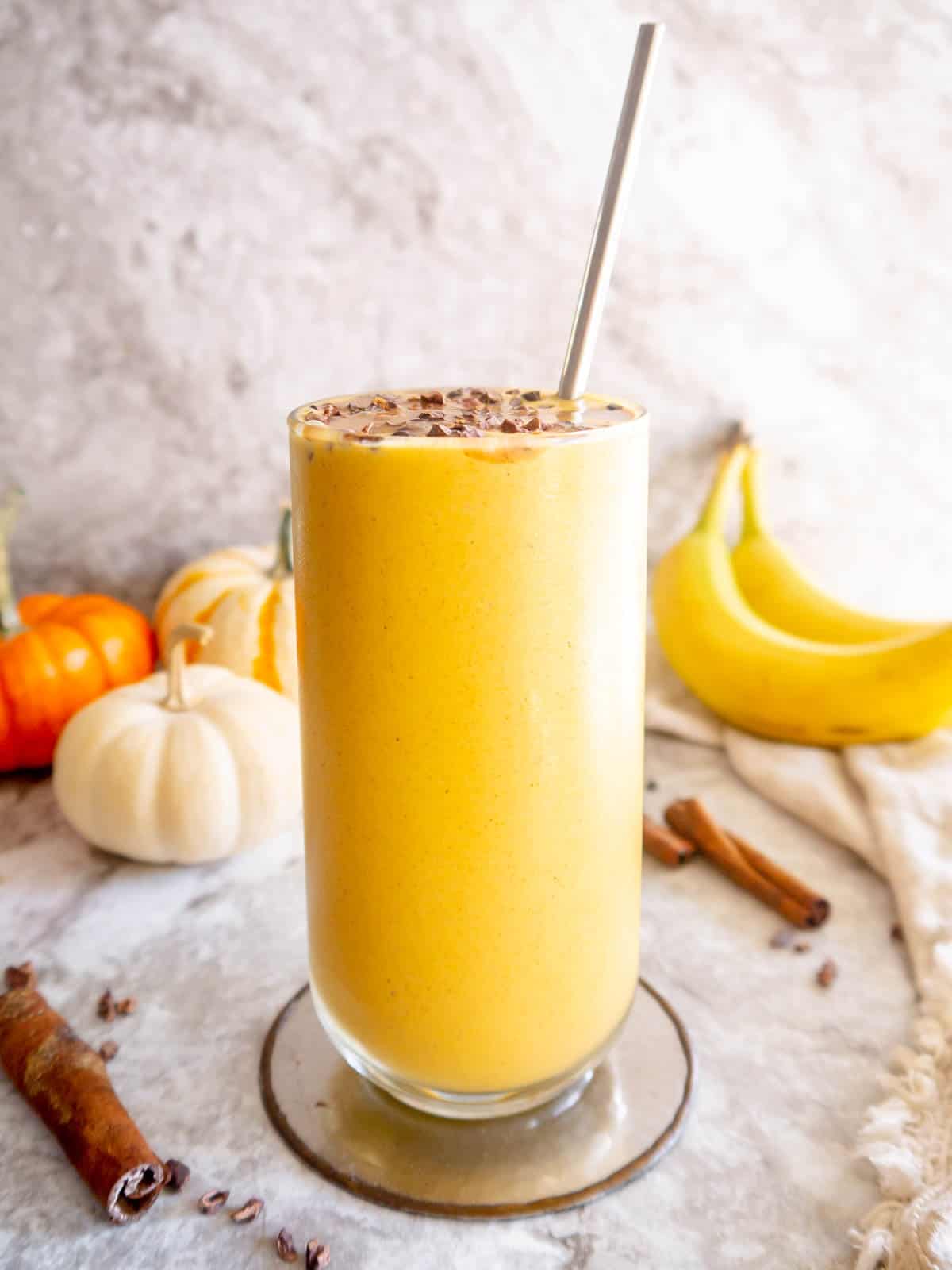Side angle of pumpkin and banana smoothie in a cup with pumpkins and bananas in the background.