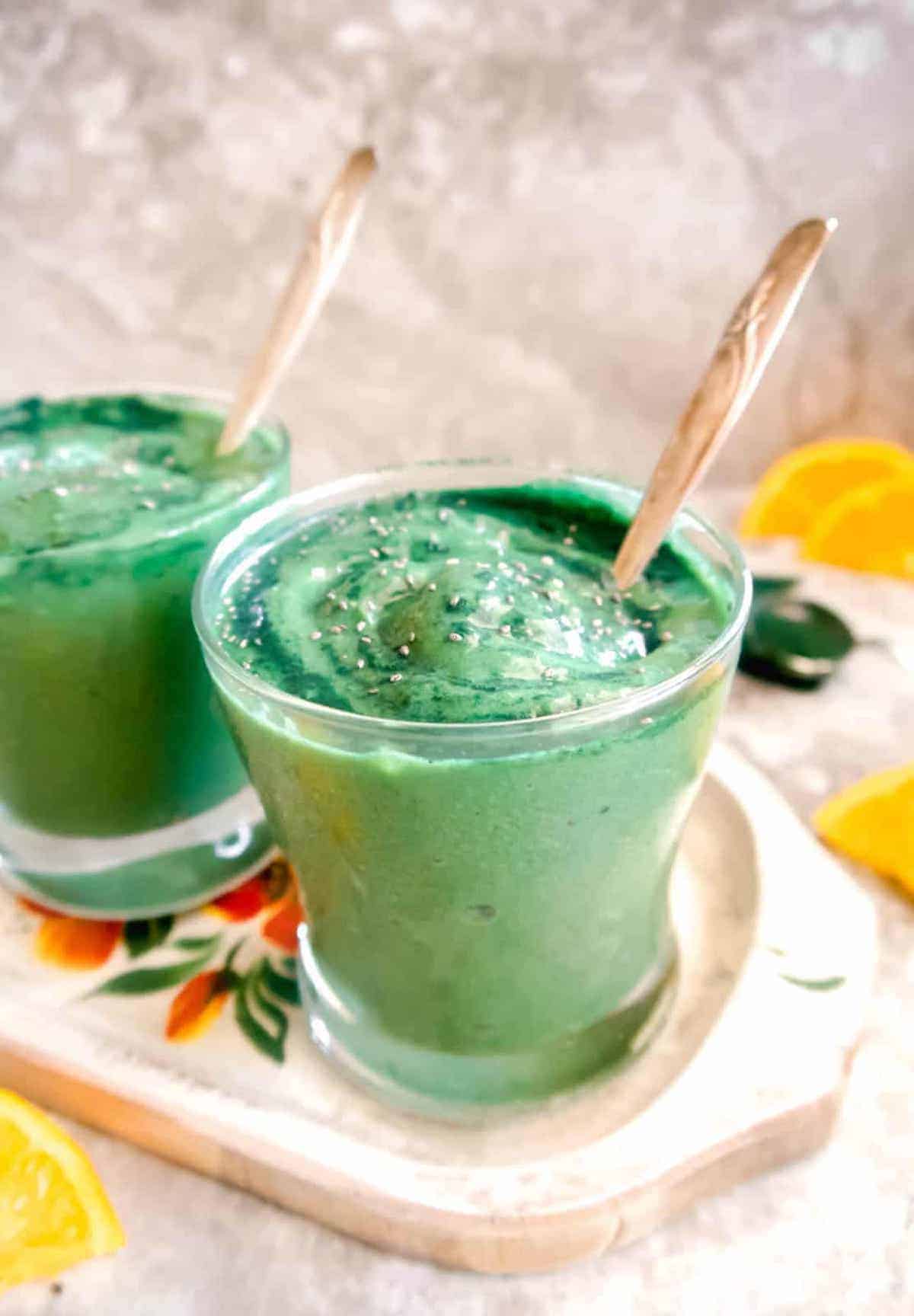 Side view of spirulina banana smoothie in two cups with oranges in the background.